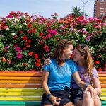 Valencia rainbow bench with colorful flowers behind and two lesbians.